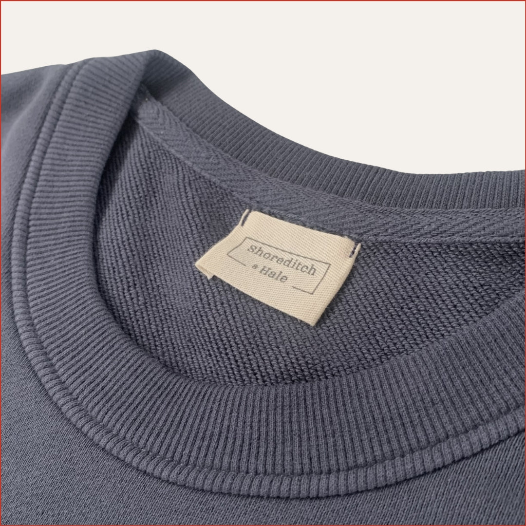 Close up image of a Carbon Blue men's sweatshirt with a twill label in the collar on a light gray background. Minimalist design casual wear by shoreditch and hale